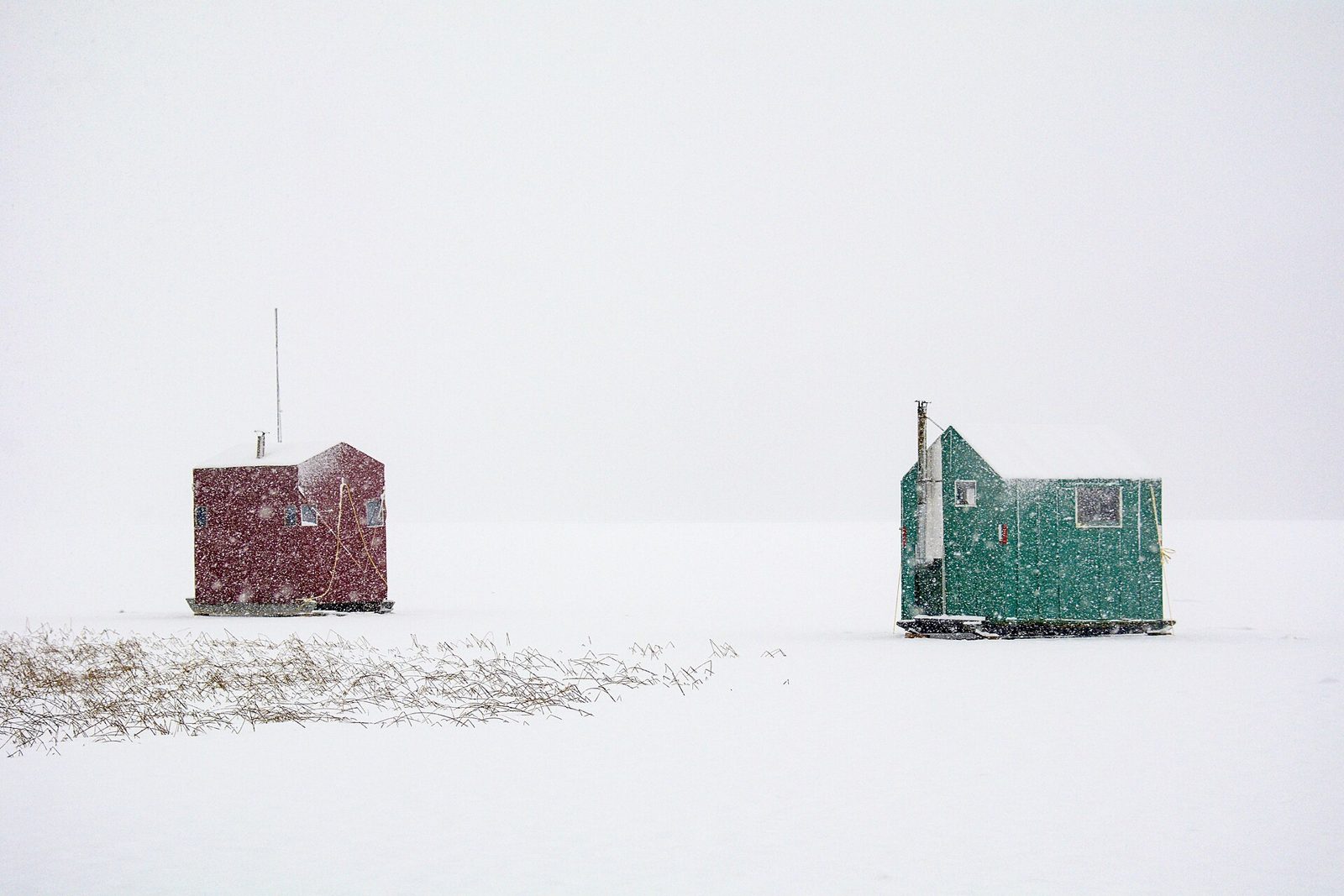 Two hard-sided ice fishing houses standing alone on a snowy frozen lake