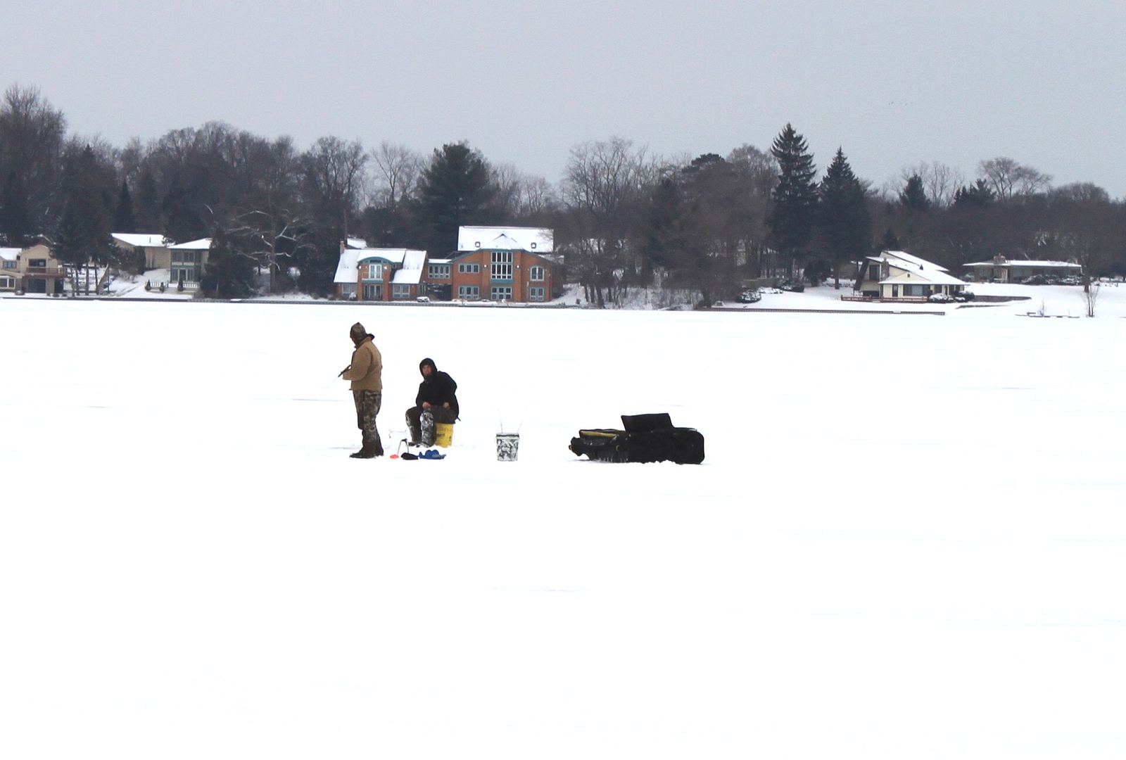 Anglers and gear sleds spread out on a frozen Michigan lake with houses along the far shore