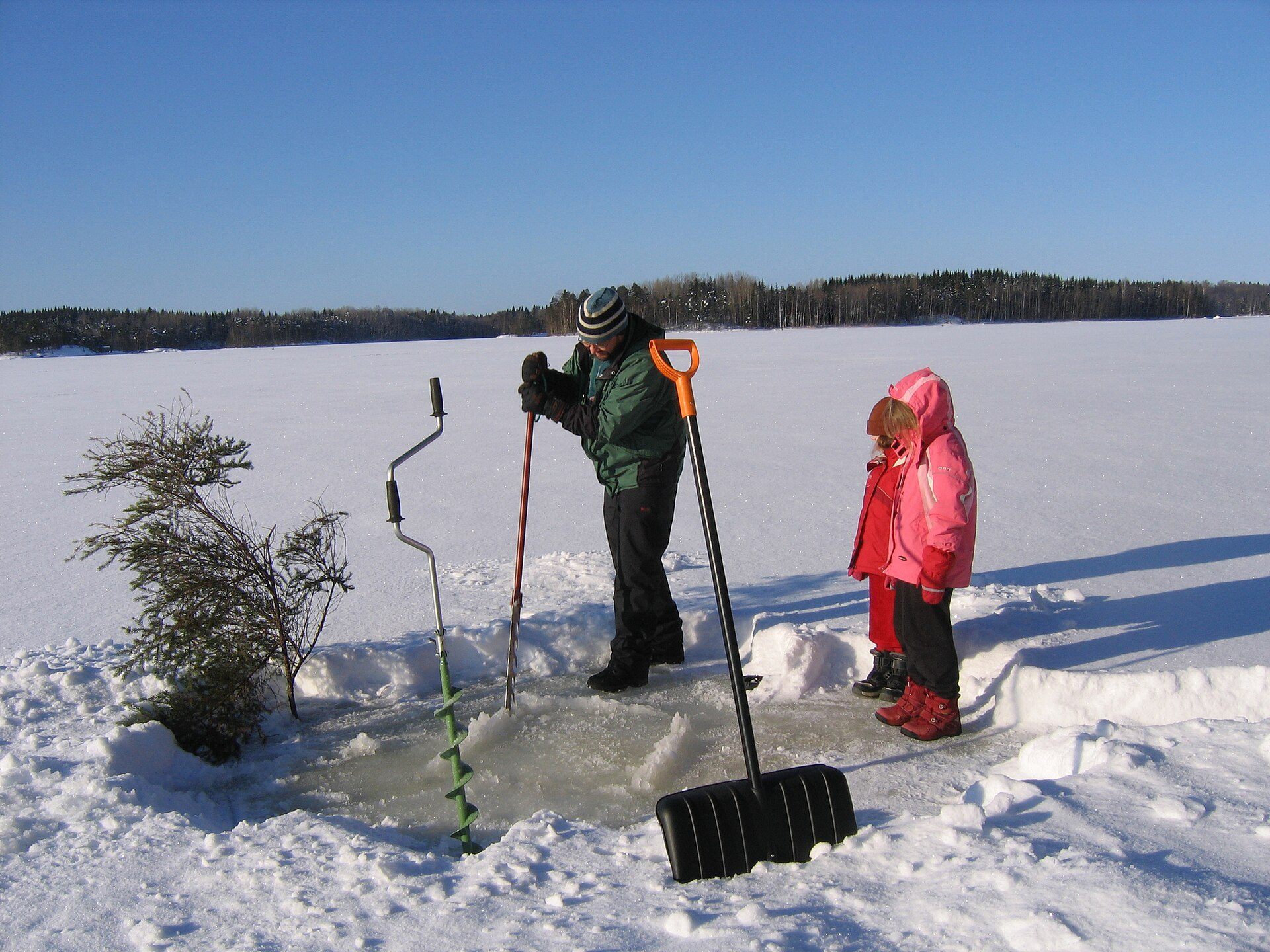 Anglers cutting a hole in thick lake ice with a hand auger and ice saw nearby