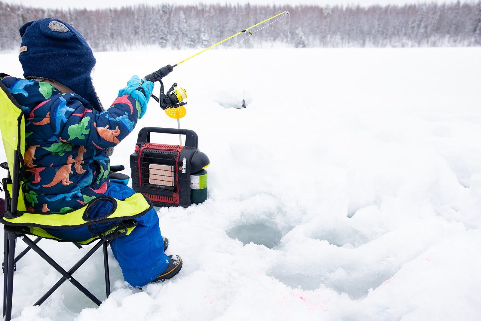 Child jigging an ice rod over a drilled hole beside a portable propane heater