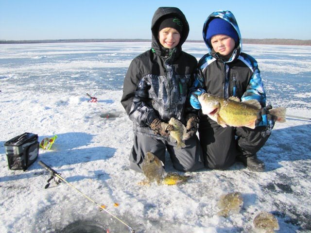 Two kids on lake ice holding panfish, with an ice rod and reel lying beside the hole