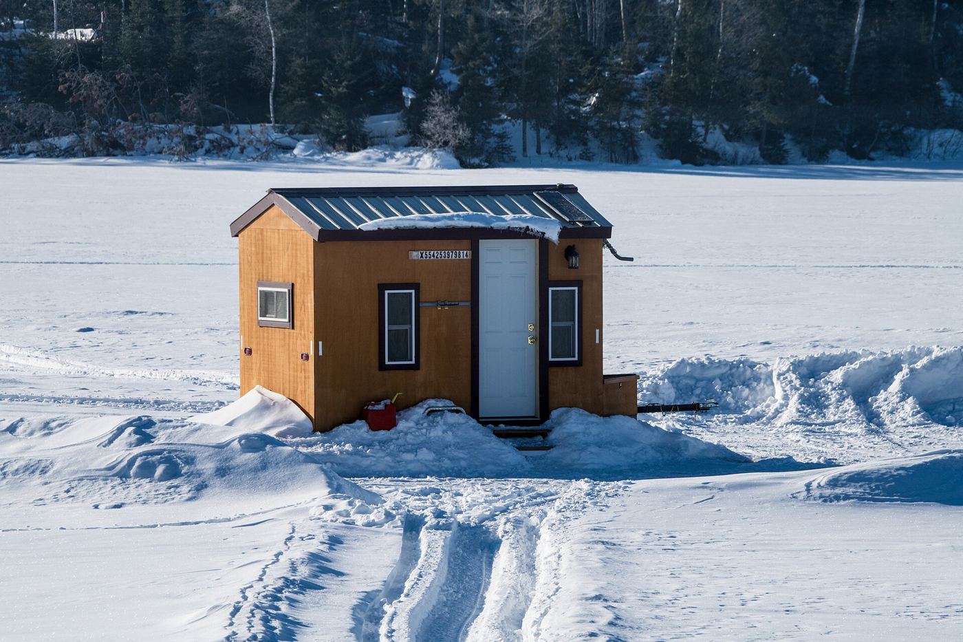 A hard-sided wooden fish house with a metal roof sitting on a snow-covered northwoods lake, with snowmobile tracks leading to it