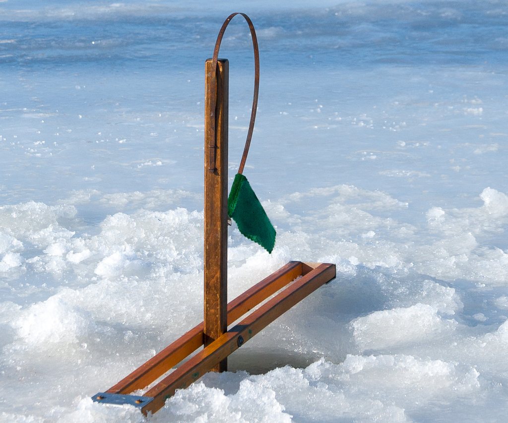 Wooden tip-up with a green flag set over a hole in lake ice, more tip-ups in the distance