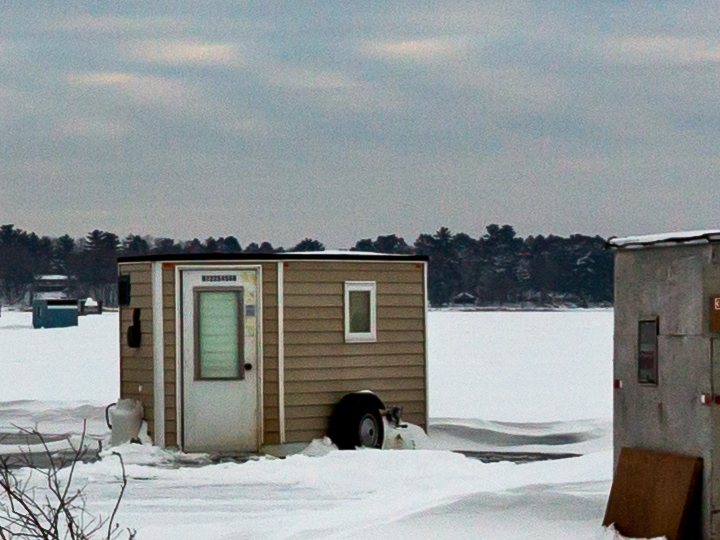 Tan sided ice fishing house on a wheeled trailer axle parked on a frozen Minnesota lake